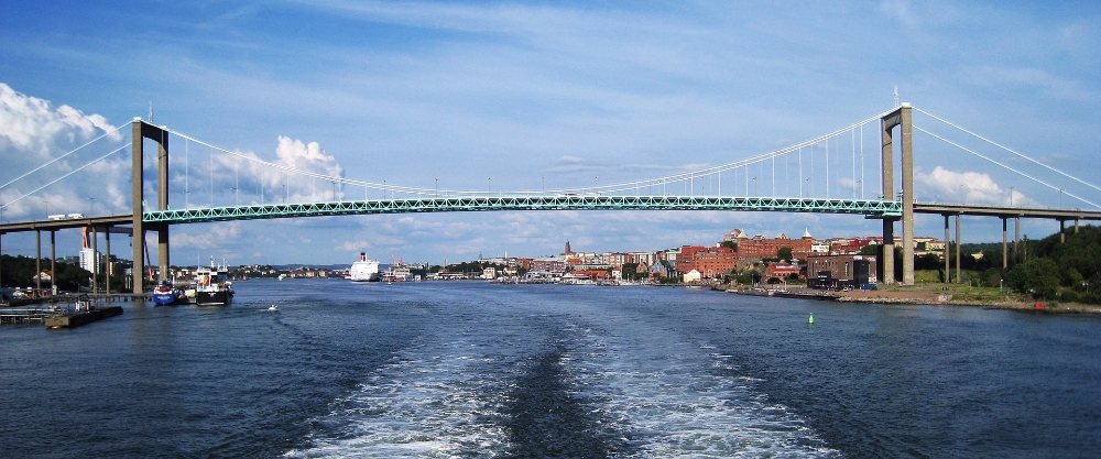 Vista del puente Älvsborg, un puente colgante verde sobre el río Göta älv, con la ciudad y barcos al fondo bajo un cielo azul con nubes blancas.